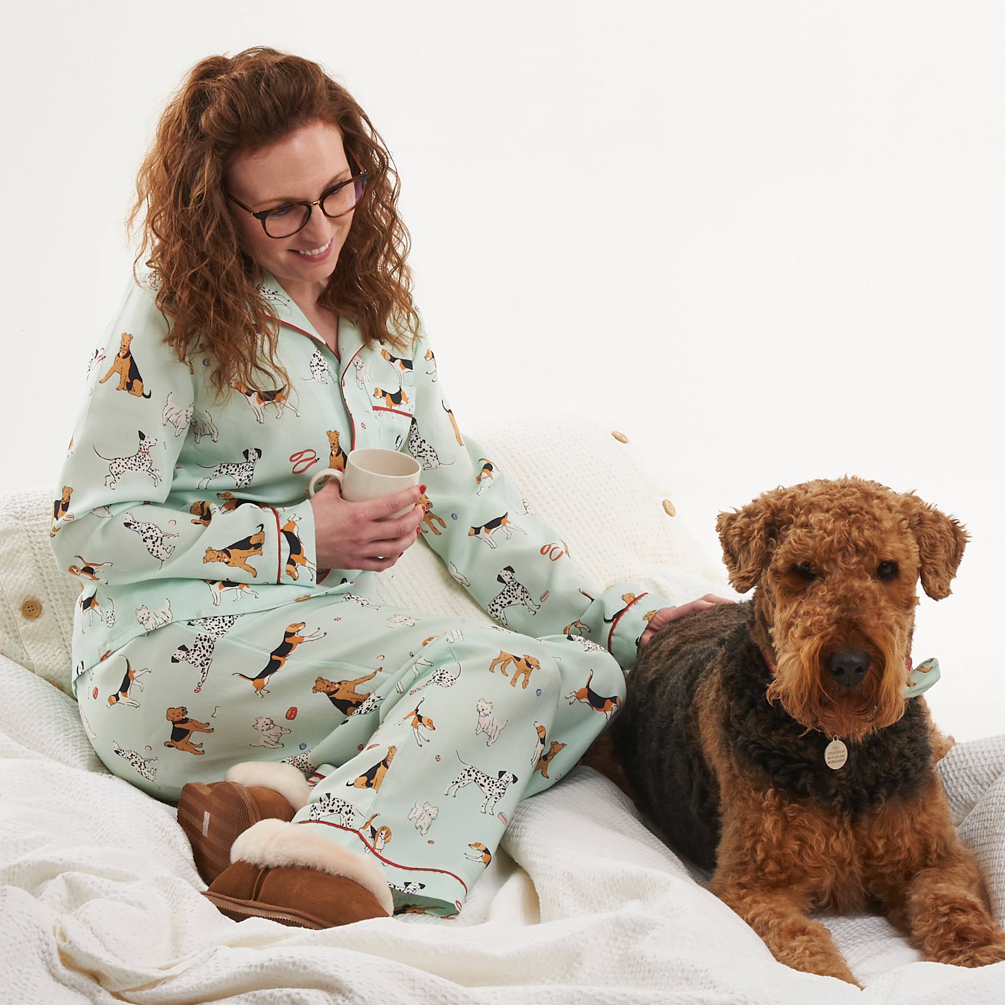 Tessie Clothing Poppy Dog Print Pyjamas Set showing a model and Airedale Terrier, sat on bedding in the pyjamas